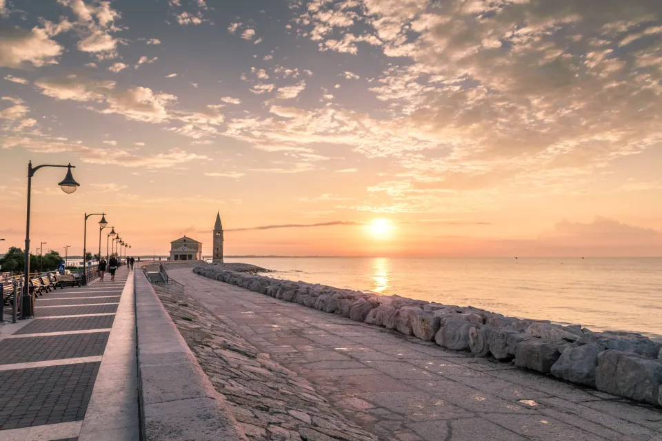 Seafront promenade of Caorle at sunset, with the Madonna dell'Angelo sanctuary in the distance